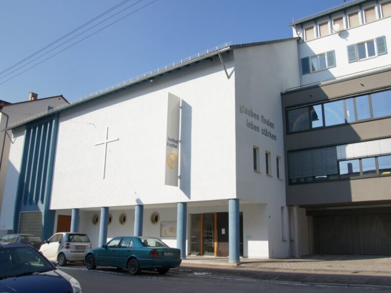 Das Gebäude der Bethelkirche von außen. Man sieht ein weißes Gebäude mit einem Kreuz außen und dem Spruch "glauben finden leben stärken".