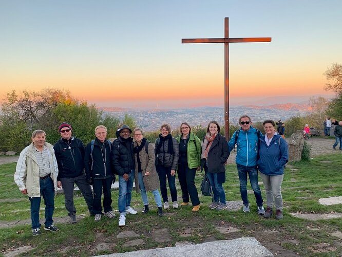 Gruppe an Menschen steht auf einem Aussichtspunkt mit Kreuz und Sonnenuntergang im Hintergrund.