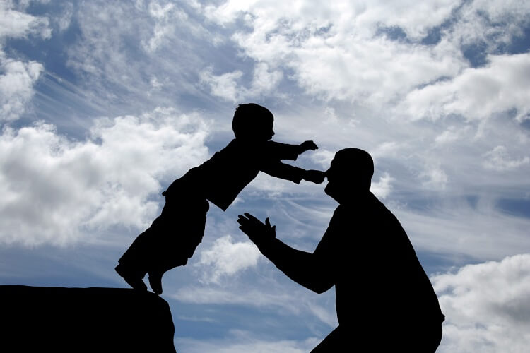 Kind springt von einem Vorsprung in die Arme von einem Erwachsenen. Im Hintergrund sieht man einen blauen Himmel, die Menschen sind nur Silhouetten.