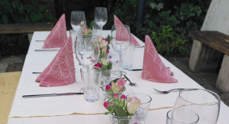 Gedeckter Tisch mit weißer Tischdecke, Rosen und rosa Stoffservietten.