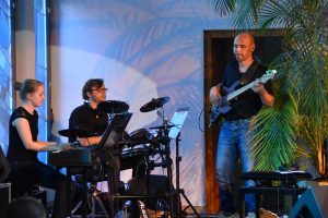 Dreiergruppe an Musikern. Eine spielt E-Piano, einer E-Schlagzeug, einer E-Gitarre.