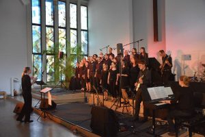 Singende Gruppe von schwarz gekleideten Menschen auf Bühne. Mit blau-rotem Licht beleuchtet.