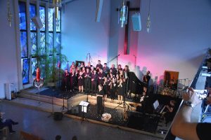 Singende Gruppe von schwarz gekleideten Menschen auf Bühne. Mit blau-rotem Licht beleuchtet.